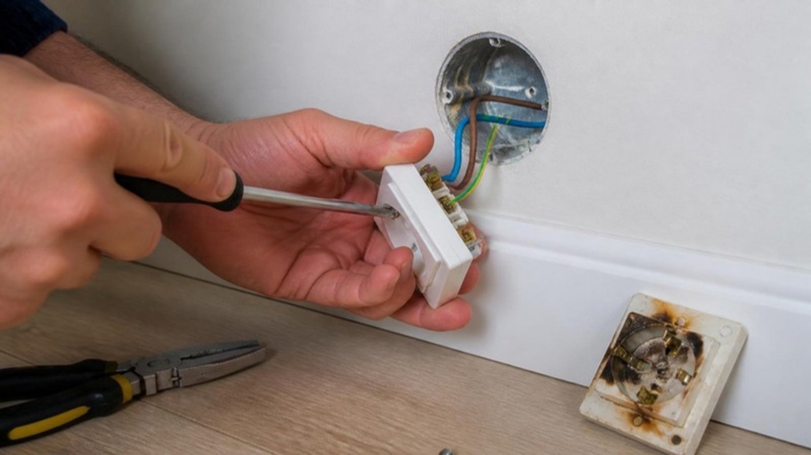 Manos de un electricista con destornillador instalando un enchufe nuevo, con el enchufe quemado retirado en el suelo